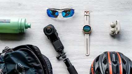 Fahrradhelm, Rucksack, Wasserflasche, Sonnenbrille, Kopfhörer, Smartwatch und eine Kamera auf einem Tisch.