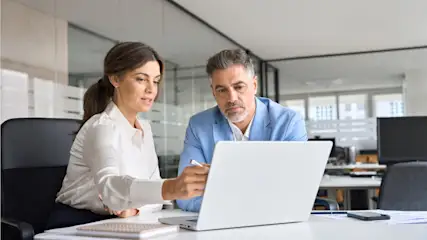 Eine Frau und ein Mann sitzen an einem Schreibtisch und schauen auf einen Laptop in einem Büro.