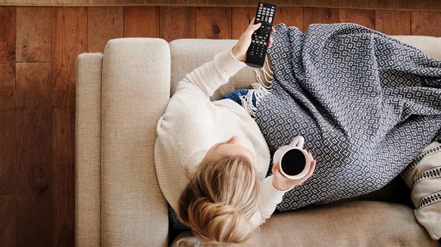 Eine Person liegt auf einem Sofa mit einer Decke, hält eine Tasse und eine Fernbedienung vor einer Holzwand.