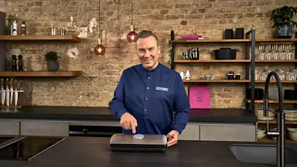 Ein Mann in blauem Hemd bedient ein silbernes Vakuumiergerät vor einer Backsteinwand mit Regalen.