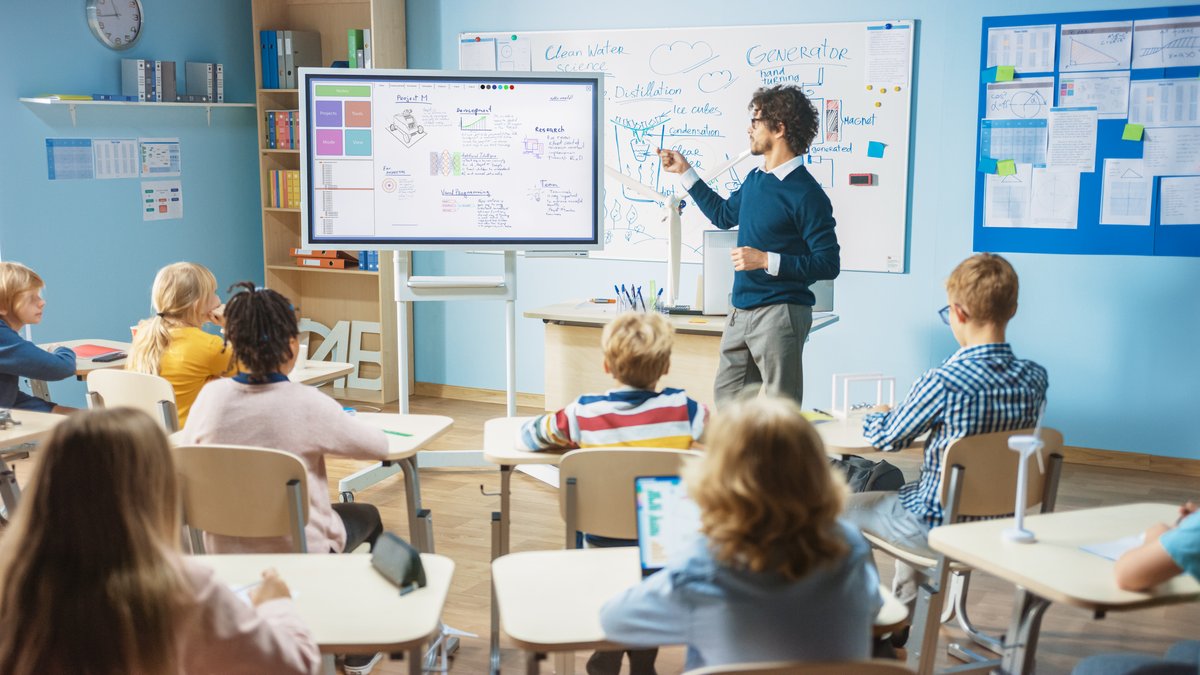 Ein Lehrer mit Brille steht vor einer Klasse und erklärt an einem Smartboard mit Diagrammen und Texten. Kinder sitzen an Tischen.