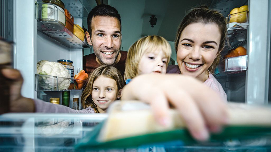 Eine Familie lächelt in einen offenen Kühlschrank, der mit Lebensmitteln wie Obst und Gemüse gefüllt ist.