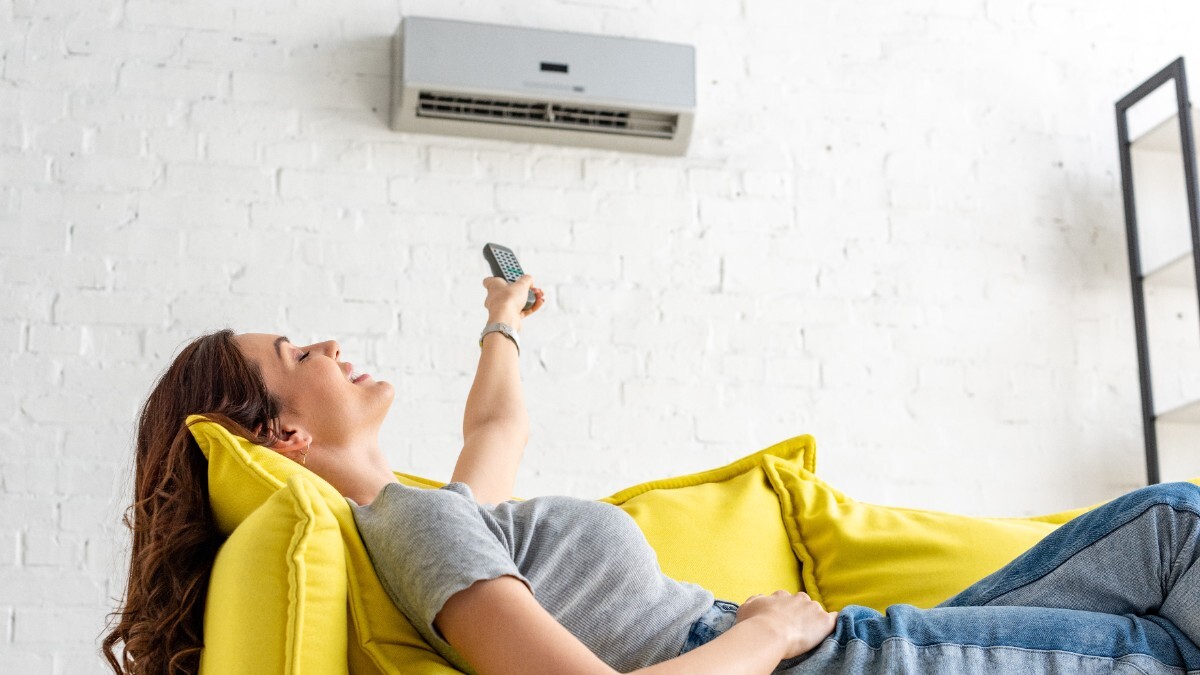 Eine Frau liegt auf dem Sofa und bedient eine Klimaanlage an der Wand mit einer Fernbedienung.