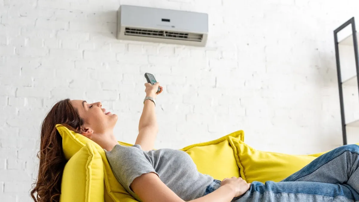 Eine Frau liegt auf dem Sofa und bedient eine Klimaanlage an der Wand mit einer Fernbedienung.