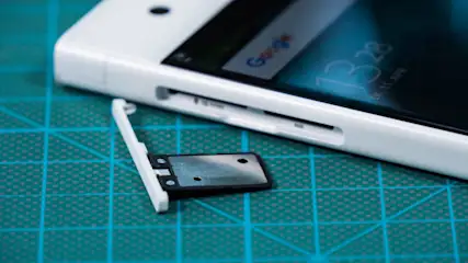 Weißes Smartphone liegt auf einer blau-grünen Gittermatte. Der SIM-Karten-Schlitten ist ausgefahren. Auf dem Display ist die Uhrzeit 13:28 zu sehen.
