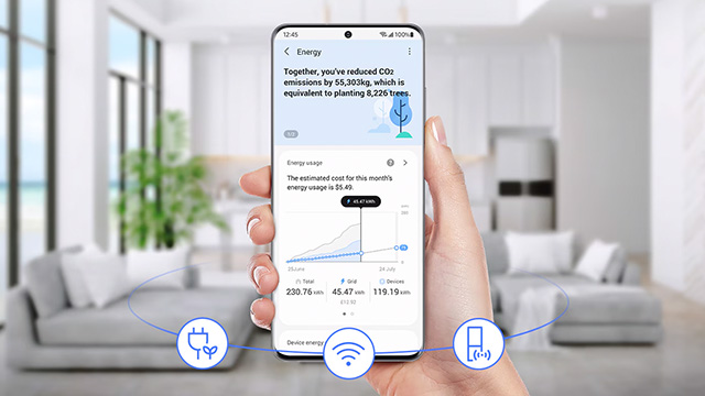 Hand hält Smartphone mit Energie-App vor Wohnzimmer. App zeigt CO2-Reduktion und Energieverbrauch an. Sofa und Pflanze im Hintergrund.