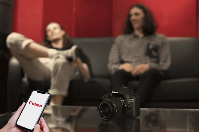 CANON EOS 2000D Kit Spiegelreflexkamera, 24,1 Megapixel, 18-55 mm Objektiv (EF-S, IS II), WLAN, Schwarz Eine schwarze Canon-Kamera steht auf einem Tisch, mit zwei Personen auf einer schwarzen Couch im Hintergrund und einem Handy in der Hand.