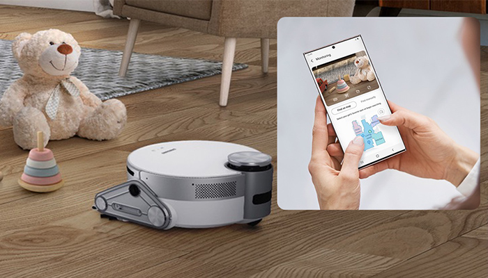 Ein Roboterstaubsauger steht auf einem Holzboden mit Spielzeug. Ein Smartphone wird in der Hand gehalten. Auf dem Bildschirm ist das gleiche Zimmer zu sehen.