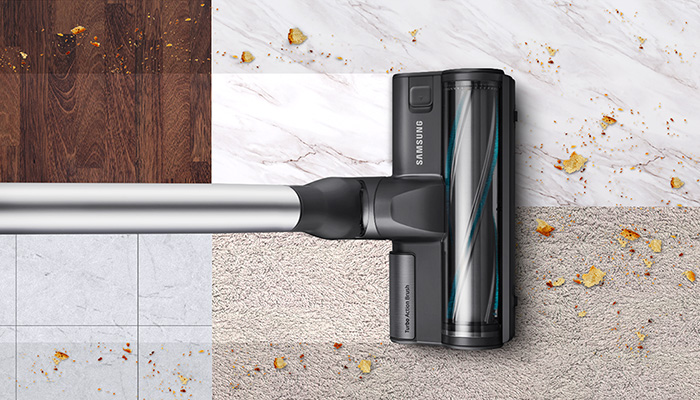 Staubsauger mit silbernem Rohr und dunkler Bürste reinigt Krümel auf Holz, Fliesen, Marmor, Teppich und rauem Untergrund.