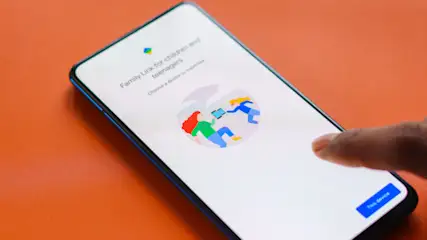 Ein Smartphone zeigt "Family Link" mit Figuren, die Geräte austauschen. Ein Finger berührt den Bildschirm über einer Schaltfläche. Der Hintergrund ist orange.