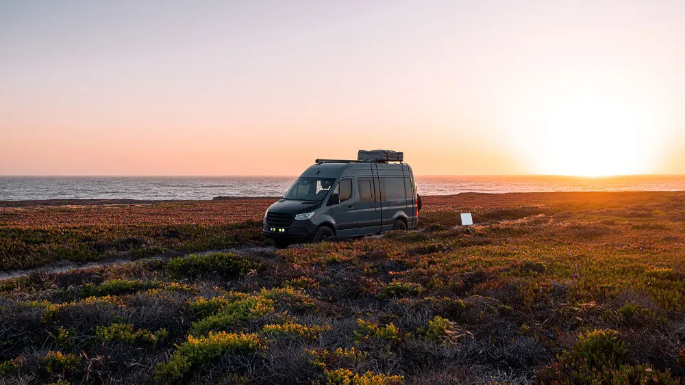 Ein grauer Van mit Dachgepäckträger steht in einem Feld mit Blick auf das Meer bei Sonnenuntergang.