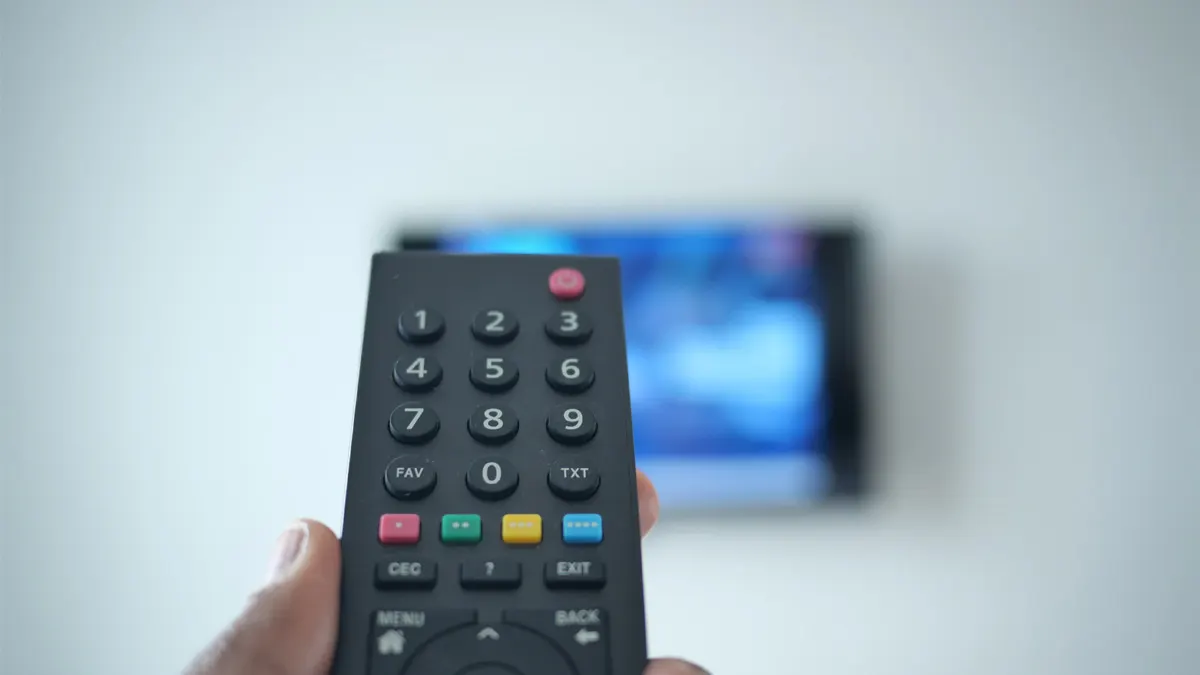 Eine Hand hält eine schwarze Fernbedienung mit bunten Knöpfen vor einem hellen Hintergrund mit verschwommenem TV.