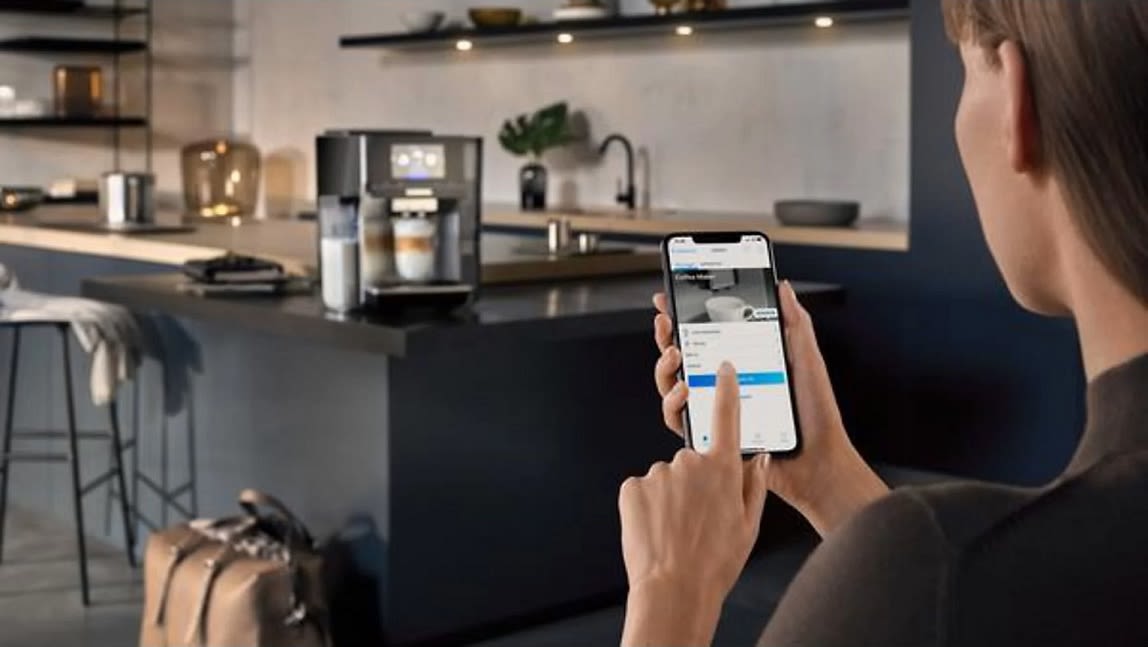 Eine Frau bedient über ihr Smartphone den Kaffeevollautomaten, welcher im Hintergrund in der Küche steht.