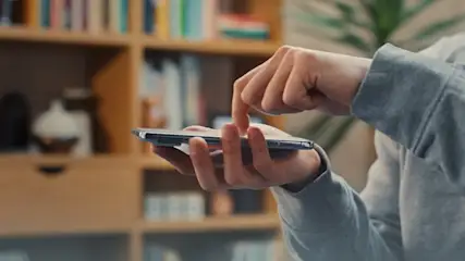 Eine Person im grauen Pullover tippt auf ein Smartphone. Im Hintergrund ein Bücherregal mit Büchern.