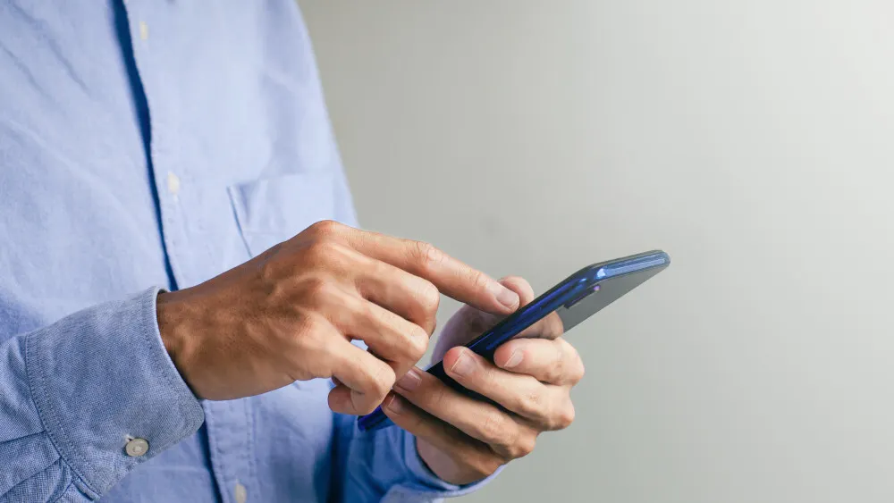 Eine Person im hellblauen Hemd bedient ein blaues Smartphone vor einem grauen Hintergrund.