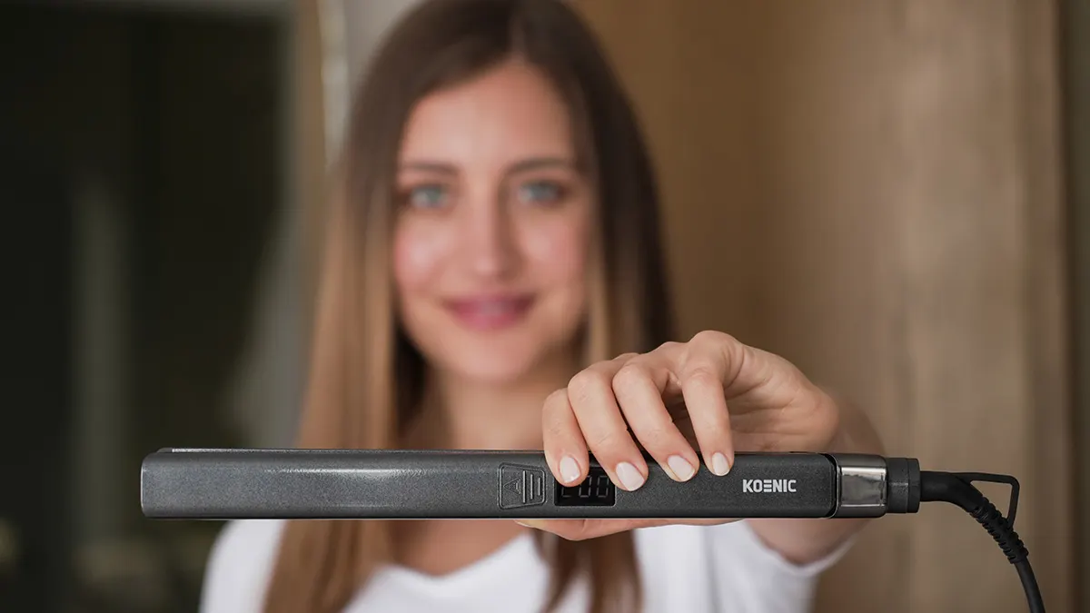 Eine junge Frau hält ein Glätteisen mit der Aufschrift KOENIC in die Kamera.