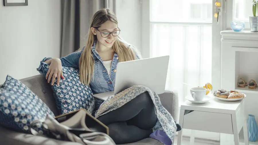 Frau mit Brille sitzt auf Couch mit Laptop. Tisch mit Tasse und Gebäck im hellen Raum.