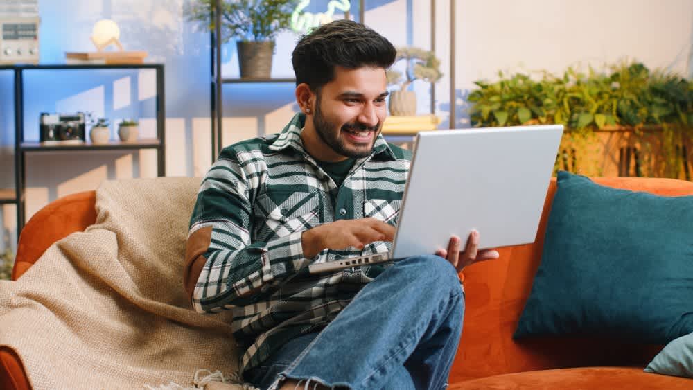 Ein Mann sitzt lächelnd, mit einem Laptop in den Händen, auf einem Sofa.