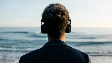 Eine Person mit Kopfhörern und einem dunklen Jackett blickt auf das blaue Meer unter einem hellen Himmel.
