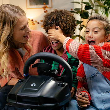 Eine Frau und zwei Kinder sitzen mit einem schwarzen Lenkrad vor einem Zimmerpflanze. Ein Kind hält dem anderen die Augen zu.