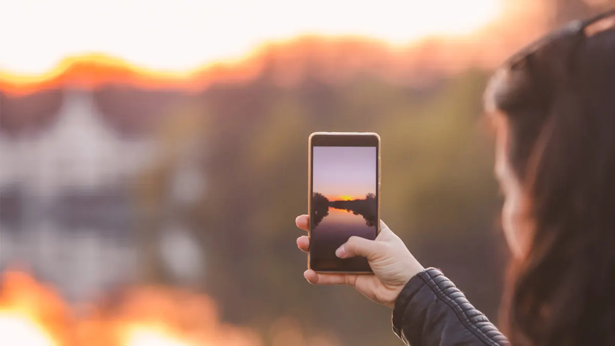 Hände halten ein Smartphone, das einen See bei Sonnenuntergang mit Bäumen im Hintergrund zeigt.