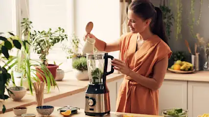 Frau gießt Flüssigkeit in Mixer mit Grünzeug in Küche mit Pflanzen, Obst und Küchenutensilien auf Arbeitsfläche.