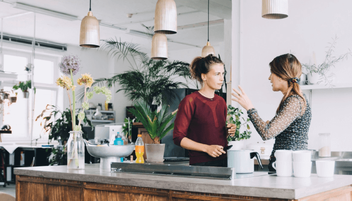 Zwei Frauen unterhalten sich in einer hellen Küche mit Pflanzen und Lampen. Auf der Arbeitsfläche stehen Tassen und eine Kochplatte.
