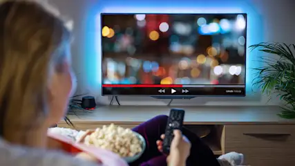 Frau sitzt mit Popcorn vor dem Fernseher und hält eine Fernbedienung in der Hand.