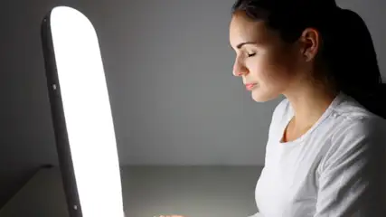 Frau in weißem Hemd mit geschlossenen Augen vor hellem Lichtpanel auf grauem Hintergrund.