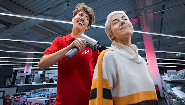 Eine Person in rotem Hemd benutzt ein Massagegerät am Nacken einer lächelnden Person im Geschäft.