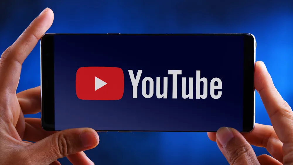 Hände halten ein Smartphone mit dem YouTube-Logo auf blauem Hintergrund. Das Logo besteht aus einem roten Rechteck mit weißem Dreieck und weißem Text.