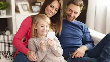 Auf einem Sofa sitzt eine Familie. Das Kind hält ein Smartphone. Die Eltern lächeln, im Hintergrund ein Regal.