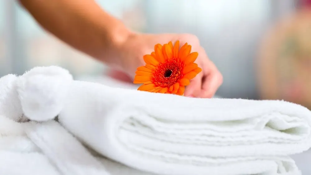 Eine Hand legt eine orangefarbene Gerbera auf einen Stapel weißer, gefalteter Handtücher.