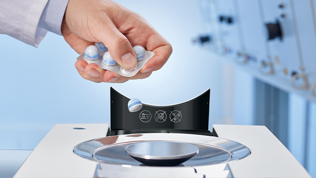 Eine Hand hält eine Blisterpackung mit runden, blau-weißen Tabletten über ein schwarzes Gerät mit einem silbernen Ring.