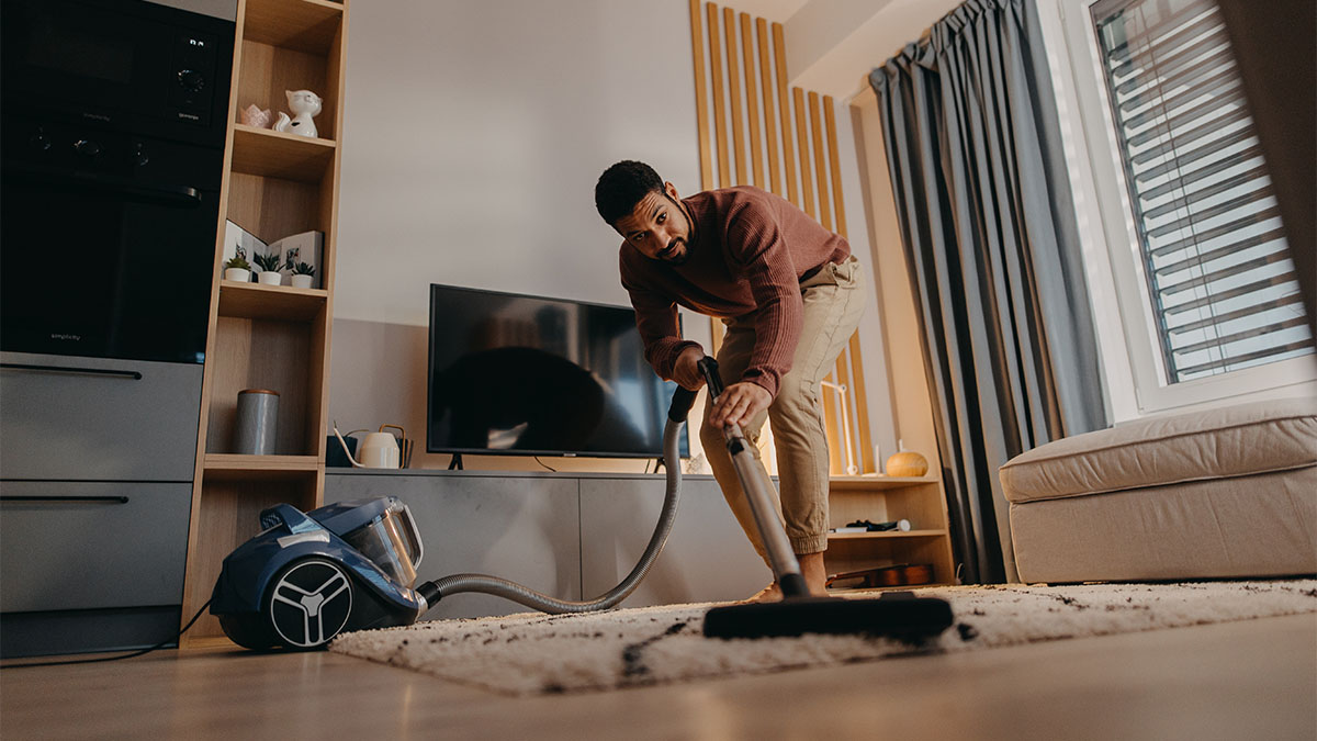 Ein Mann saugt im Wohnzimmer einen hellen Teppich mit einem blauen Bodenstaubsauger. (Adobe Stock, Datei Nr. 498271480)