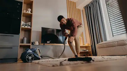 Ein Mann saugt im Wohnzimmer einen hellen Teppich mit einem blauen Bodenstaubsauger. (Adobe Stock, Datei Nr. 498271480)