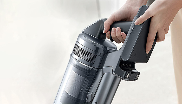 Eine Person hält einen grauen, kabellosen Staubsauger mit durchsichtigem Staubbehälter und schwarzem Griff.