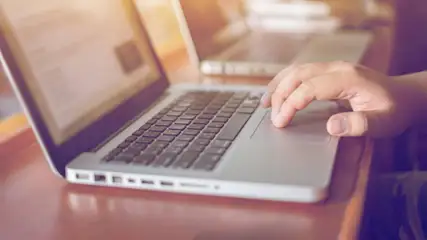 Eine Hand bedient das Touchpad eines silbernen Laptops auf einem Holztisch. Ein zweiter Laptop ist im Hintergrund teilweise sichtbar.