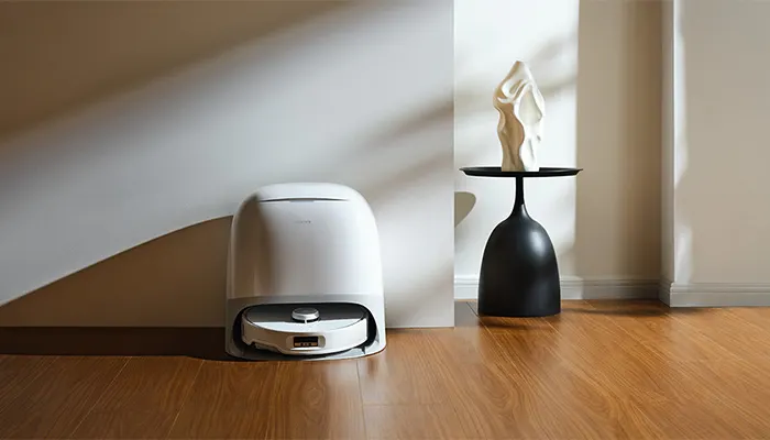 Ein weißer Roboterstaubsauger steht in einer Ladestation neben einem schwarzen Beistelltisch mit einer weißen Skulptur.