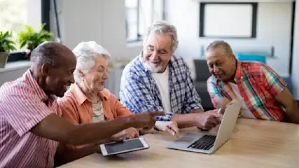Vier Senioren sitzen am Tisch und schauen auf einen Laptop. Einer der Männer zeigt auf den Bildschirm. Ein Tablet liegt daneben.