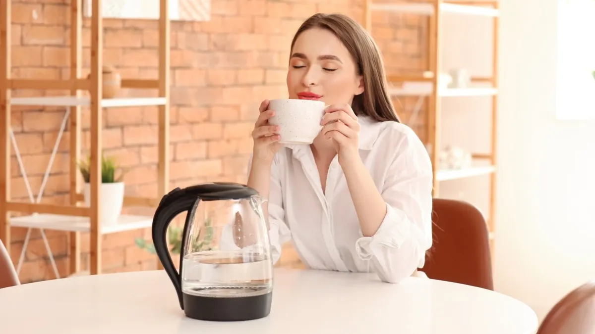 Eine Frau sitzt an einem Tisch und trinkt aus einer Tasse. Vor ihr steht ein Wasserkocher.