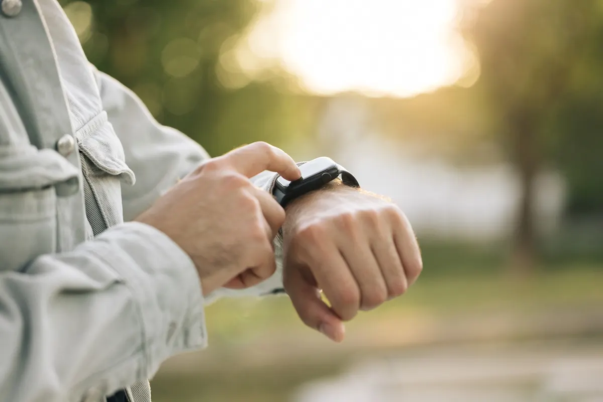 Eine Person mit hellgrauer Jacke bedient eine schwarze Smartwatch am Handgelenk vor grünem Hintergrund.