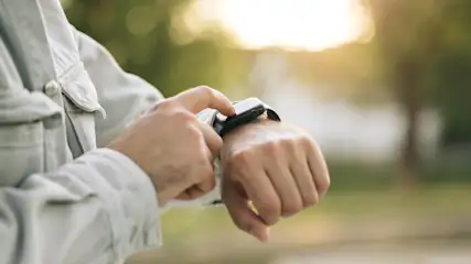 Eine Person mit hellgrauer Jacke bedient eine schwarze Smartwatch am Handgelenk vor grünem Hintergrund.