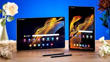 Zwei Tablets auf einem Tisch mit Stiften. Auf den Bildschirmen sind App-Symbole und ein gelbes Muster zu sehen. Blumen im Hintergrund.