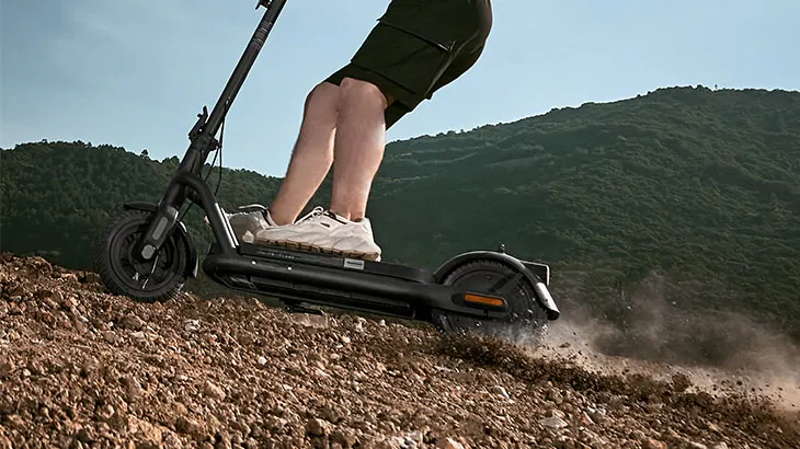 Eine Person fährt einen schwarzen Elektroroller auf einem unbefestigten Hügel mit einem bewaldeten Berg im Hintergrund.