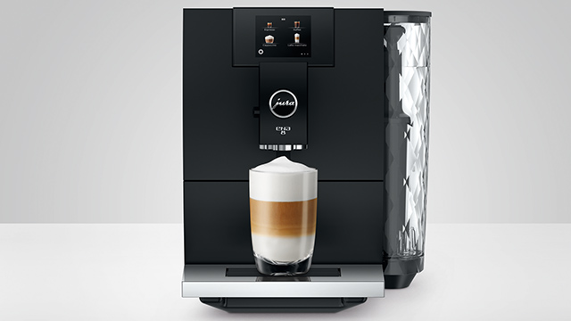 Eine Kaffeemaschine in Schwarz mit einem transparenten Wassertank und einem Latte Macchiato-Glas auf einer silbernen Ablage.