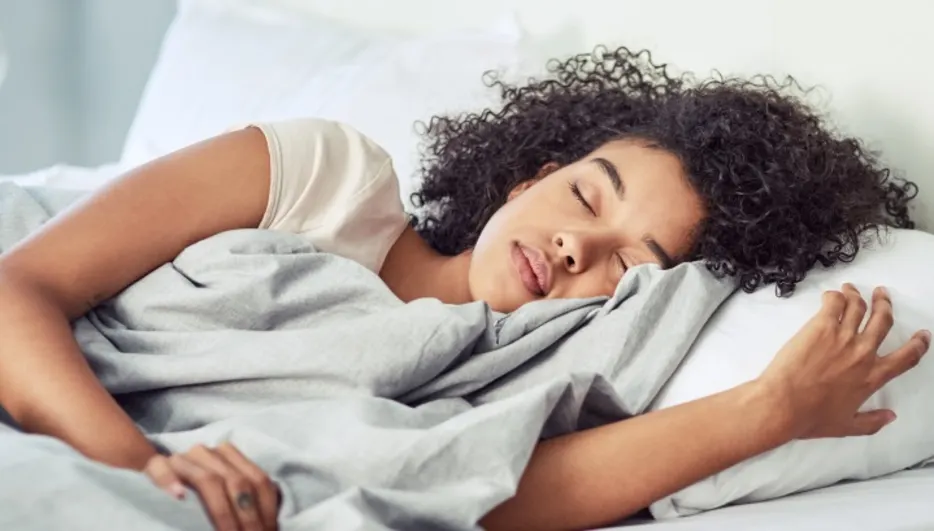 Eine Person schläft in einem Bett unter einer grauen Decke, mit einem weißen Kissen unter dem Kopf.