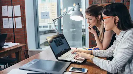 Zwei Frauen an einem Tisch mit Laptop, Grafiktablett und Smartphone. Eine Schreibtischlampe leuchtet. Ziegelwand und Fenster im Hintergrund.