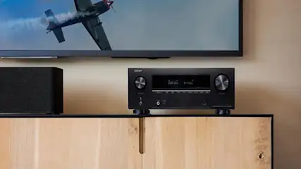Auf einem Holzschrank steht ein schwarzer Denon-Verstärker unter einem Fernseher mit einem Flugzeug darauf. Links steht ein schwarzer Lautsprecher.