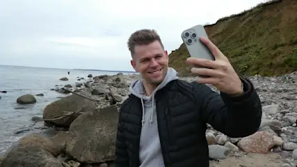 Ein Mann in schwarzer Jacke macht ein Selfie mit einem grauen Smartphone am steinigen Strand. Im Hintergrund sind Meer und Klippen.
